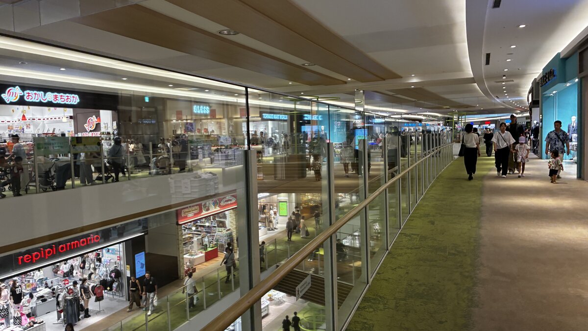 A wide, mostly empty indoor corridor inside AEON Kita-Nagoya connecting multiple floors, with glass railings, green carpet, a repipi armario children's clothing shop and an Okashi no Machioka candy shop visible on the level below, and a few families including a parent with a stroller walking through