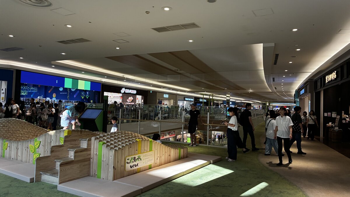 A busy indoor walkway inside AEON Kita-Nagoya mall in Aichi Prefecture, with families and shoppers passing a wooden children's play structure called Kodomoen Mokumoku and capsule-toy machines visible in the background
