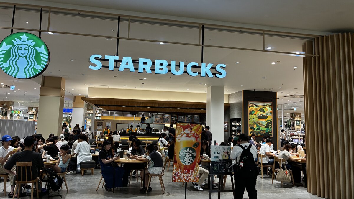 A busy Starbucks storefront inside AEON Kita-Nagoya, moderately crowded seating area, with a U24 student-discount promotional sign and orange-drink seasonal signage visible near the counter