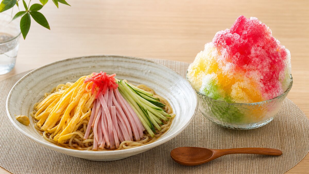 A chilled bowl of hiyashi chuka cold ramen noodles topped with thin egg strips, cucumber, ham, and tomato, next to a glass of iced mugicha barley tea on a wooden table