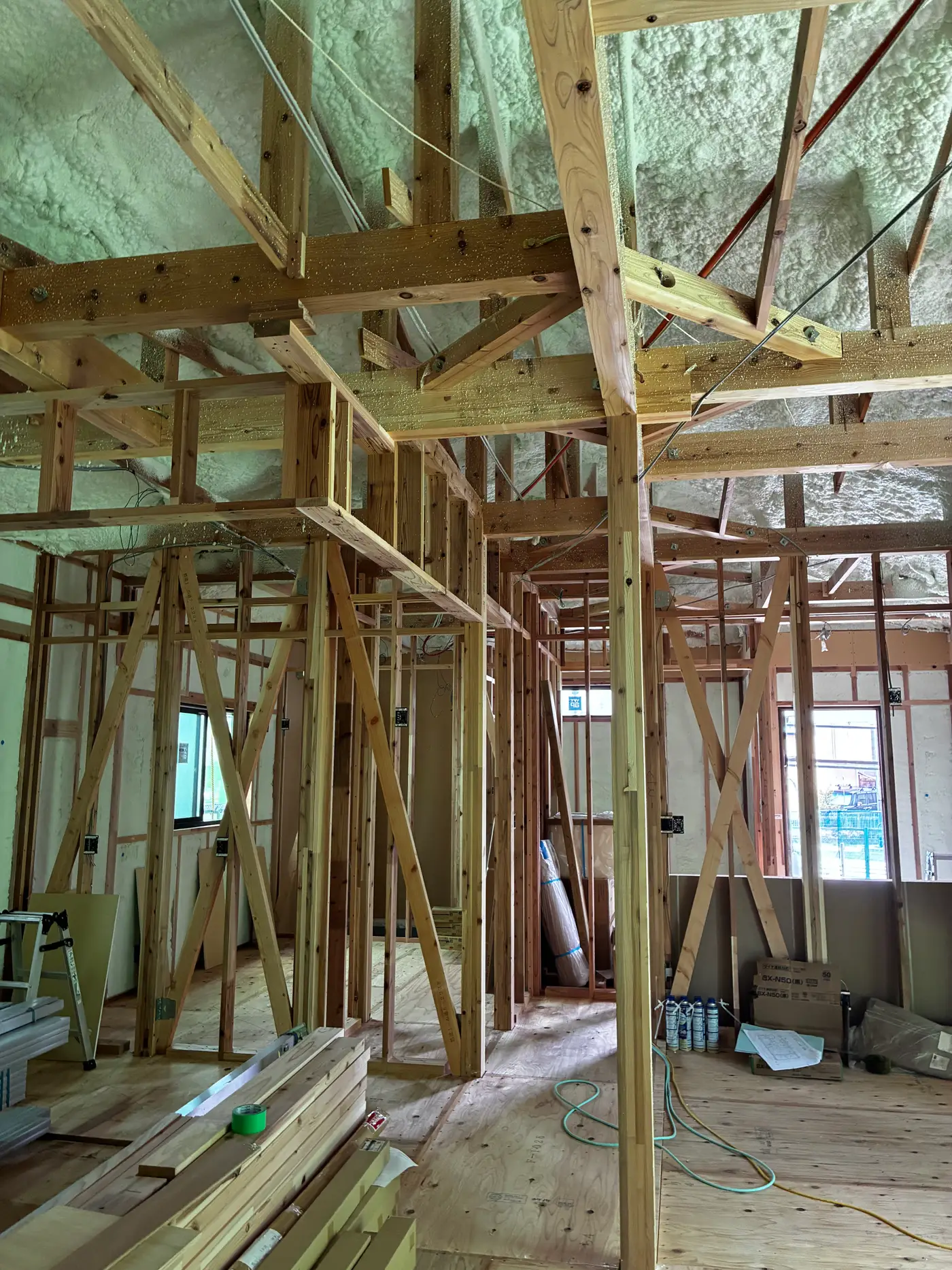 Post-and-beam frame of a Japanese house viewed from below, showing wooden beams, posts, diagonal braces, and sprayed foam insulation on the wall behind