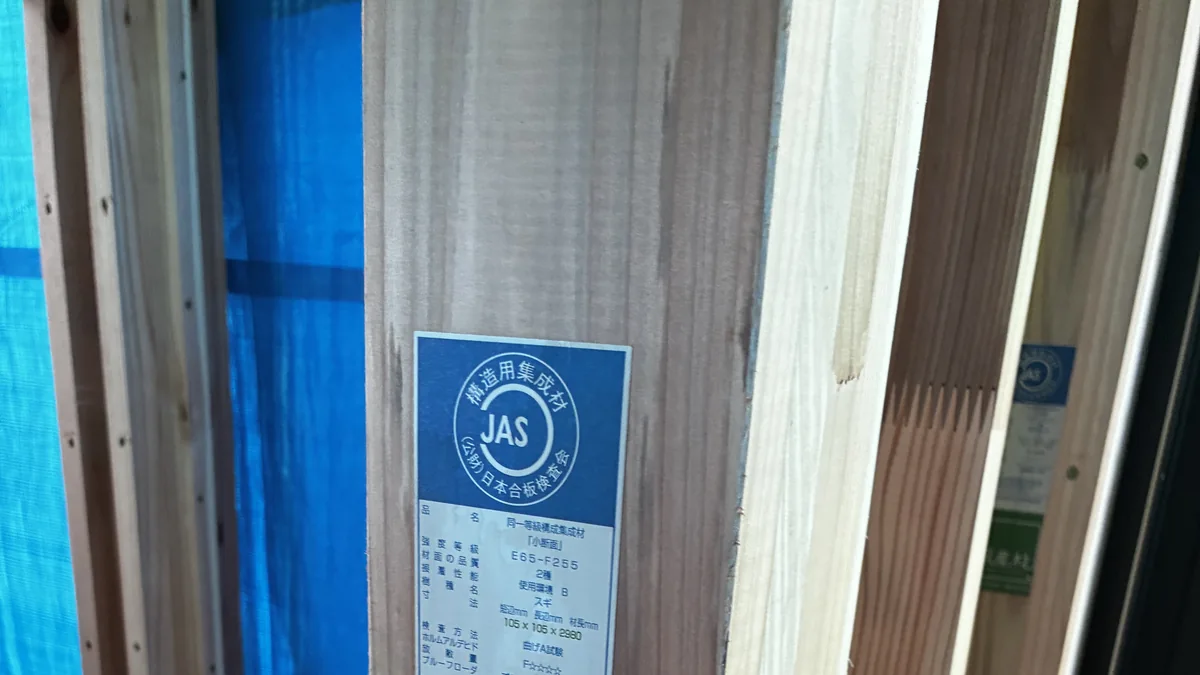 A JAS certification label on a structural glulam post in a Japanese house under construction, showing strength grade E65-F255, dimensions 105 by 105 by 2980 millimeters, and a 100 percent domestic timber mark
