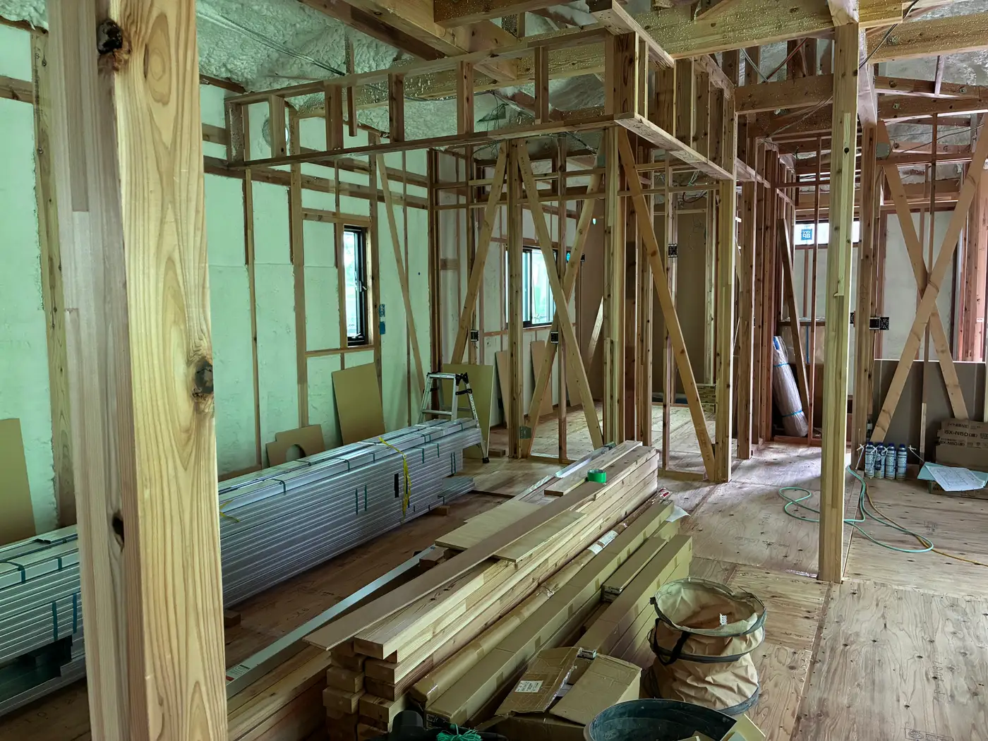 Open living and dining area of a Japanese house still at bare frame stage, with construction materials stacked on the plywood floor deck
