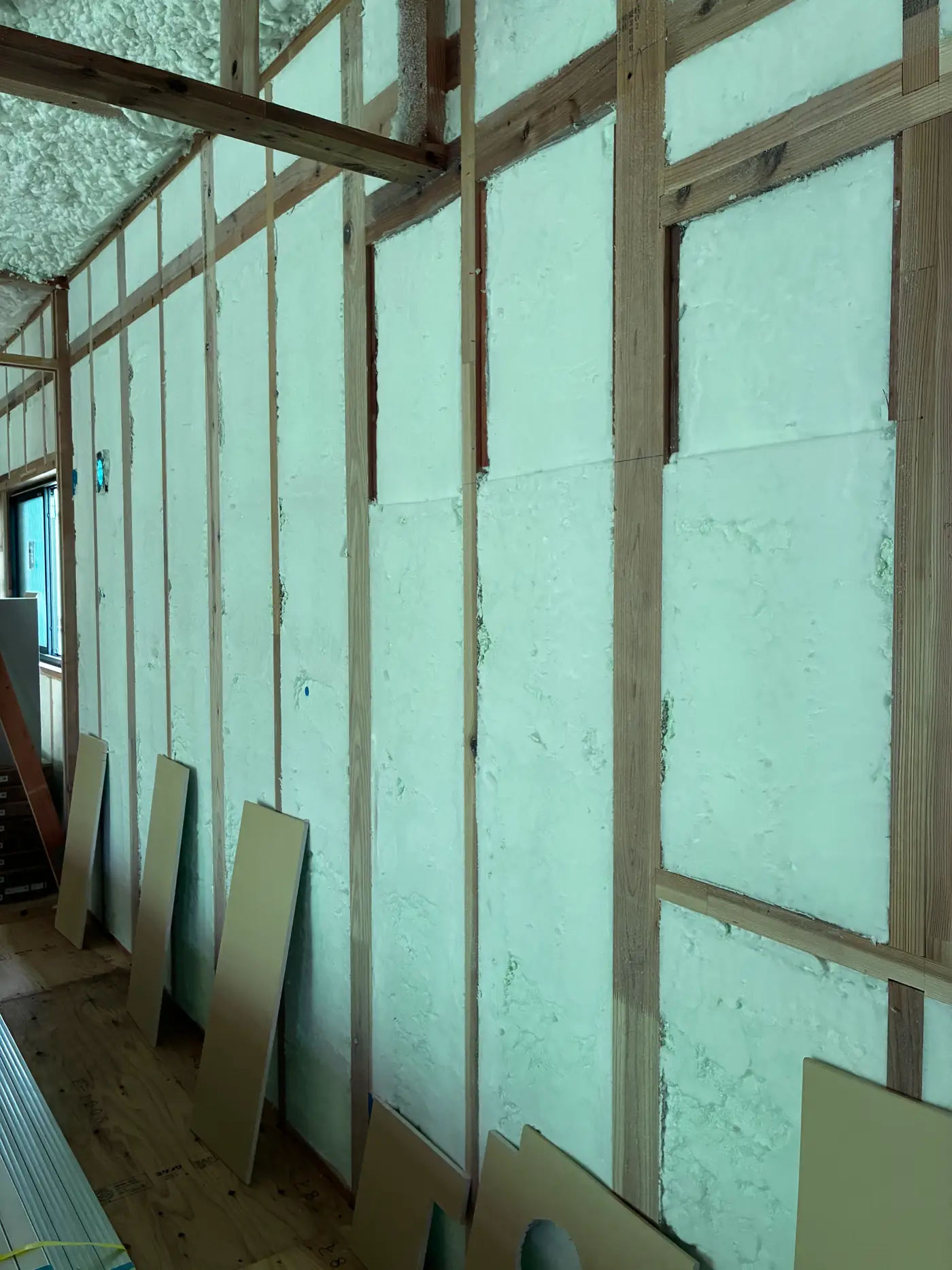 Close view of foam-insulated ceiling and roof plane with wooden ceiling battens installed below it and plasterboard sheets stacked ready to be hung