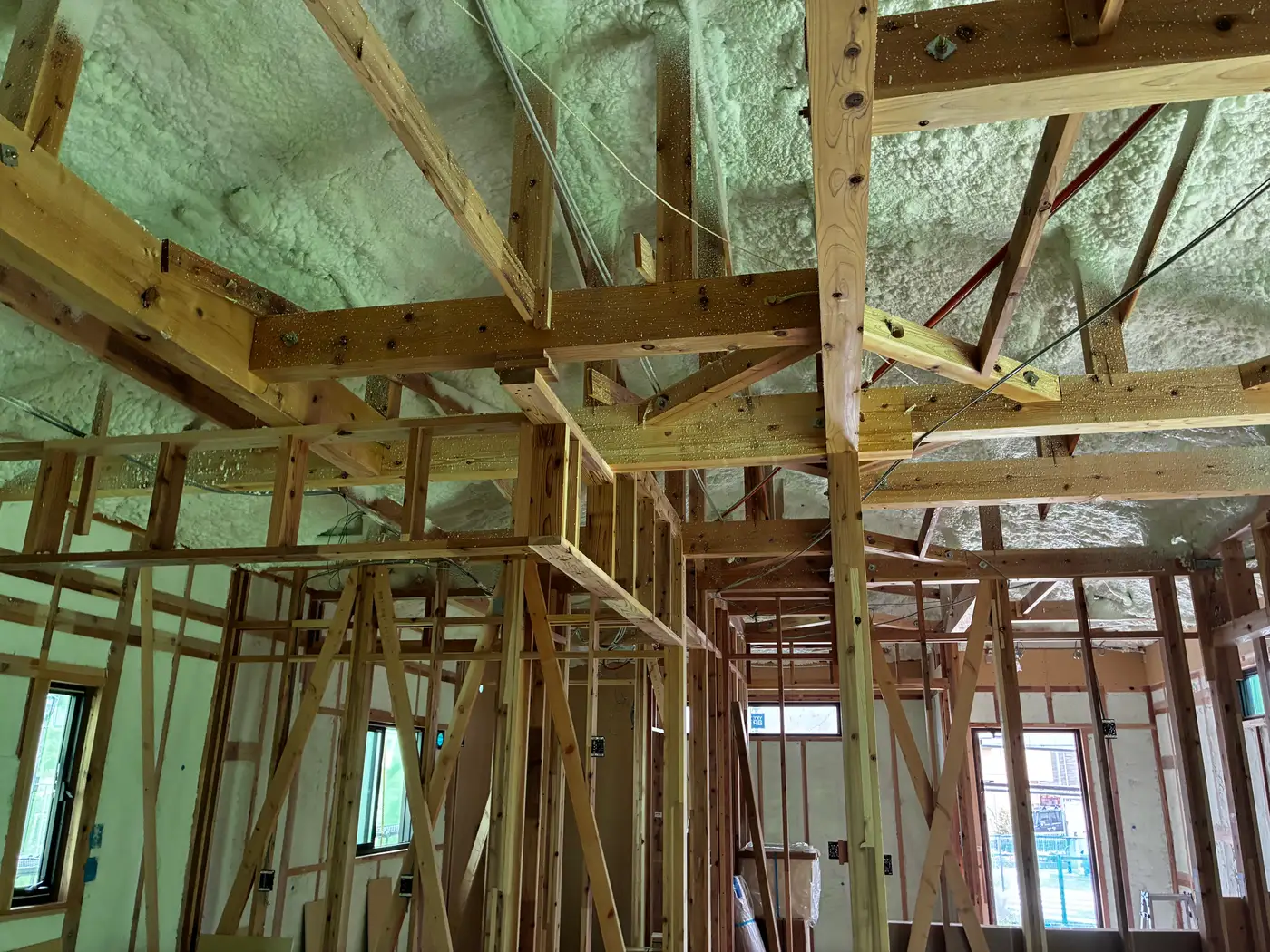 Looking up into the roof frame of a Japanese house under construction, with sprayed rigid foam insulation covering the roof plane between exposed beams and rafters