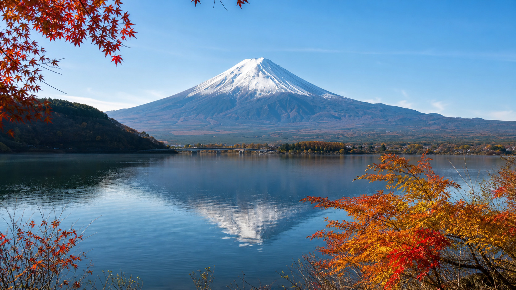 Snow-capped Mount Fuji rising above a lake with autumn-colored trees in the foreground under a clear blue sky