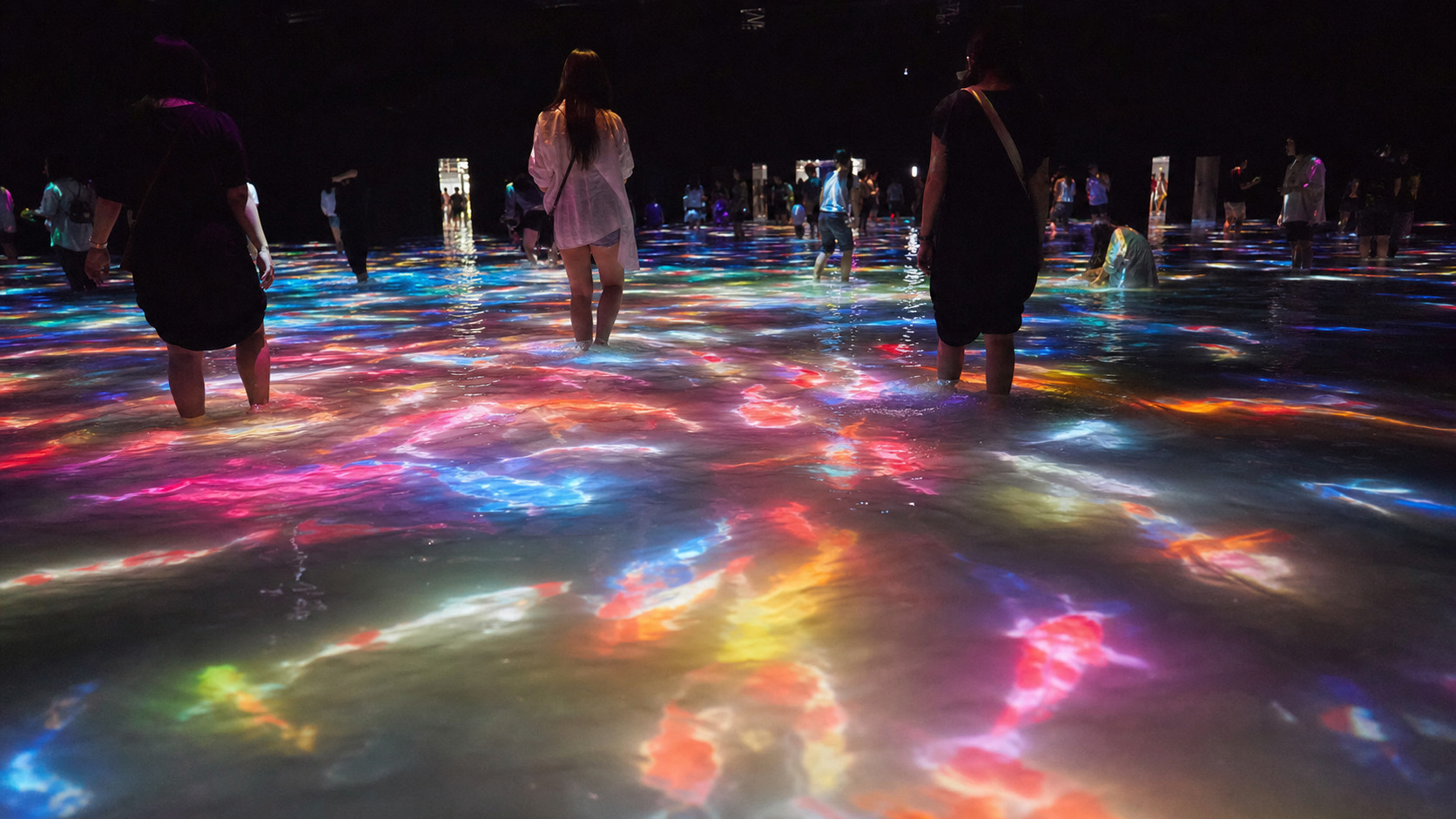 Visitors wading barefoot through a shallow reflective pool of water illuminated by colorful projected koi fish light installations at teamLab Planets Tokyo